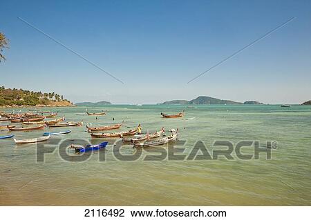Stock Image - Thailand, Phuket, Rawaii Beach, Longtail boats along the shoreline, Land in distance.. Fotosearch