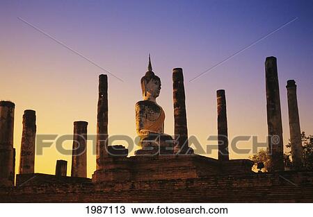 Stock Image - Thailand, Sukhothai, Wat Mahathat, Buddha Statue With Many Pillars At Sunset, Blue And Orange Sky.. Fotosearch