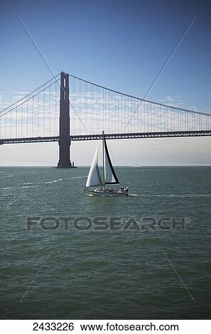 Stock Photograph - The Golden Gate Bridge is seen behind a sailboat in San Francisco Bay; San Francisco, California, United States of America. Fotosearch