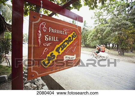 The Parrilla Grill, Las Piedras Restaurant Sign Hanging And Surfers Ride Past On Their Bicycles In Santa Teresa And Mal Pais (Malpais) On The Nicoya Peninsula; Puntarenas Province, Costa Rica View Large Photo Image Picture - The Parrilla Grill, Las Piedras Restaurant Sign Hanging And Surfers Ride Past On Their Bicycles In Santa Teresa And Mal Pais (Malpais) On The Nicoya Peninsula; Puntarenas Province, Costa Rica. Fotosearch