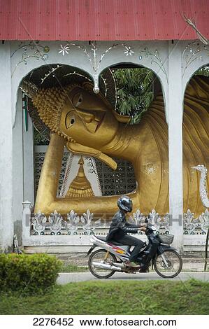 Stock Image - The reclining Buddha at Wat Somdet appearing to watch the traffic flow past; Sangkhlaburi, Kanchanaburi, Thailand. Fotosearch