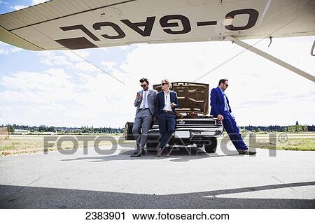 Stock Image - Three businessmen leaning against a car with an open trunk and an airplane; Langley, British Columbia, Canada. Fotosearch