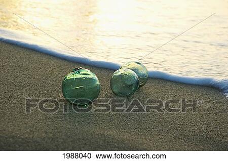 Stock Image - Three Glass Fishing Floats Rest On The Sandy Shoreline. Fotosearch