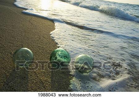 Stock Image - Three Glass Fishing Floats Roll On The Sandy Shoreline With Ripples Of Water And Seafoam. Fotosearch