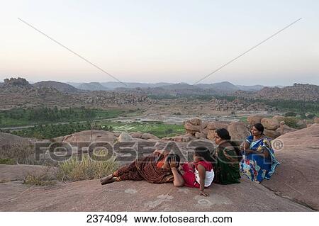 Picture - Three women and a girl sit on a rock surface overlooking the landscape; Hampi, Karnataka, India. Fotosearch