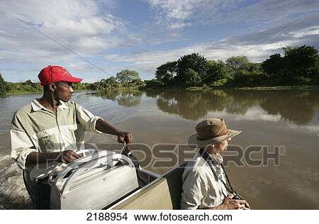 Picture - Tourist and tour guide ride in a boat down a tranquil river, Lower Zambezi National Park; Zambia. Fotosearch