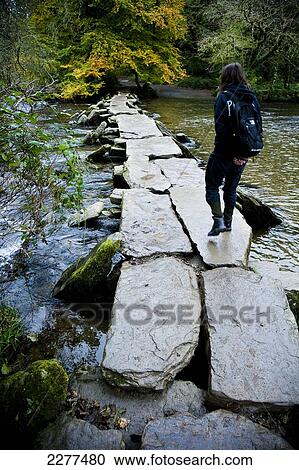 Stock Image - Tourist crossing The Tarr Steps, a prehistoric clapper bridge across the River Barle, Exmoor National Park; Somerset, England. Fotosearch