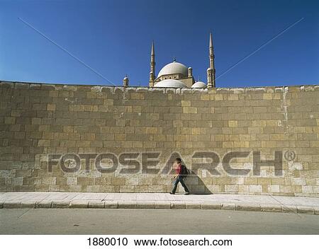 Stock Image - Tourist Walking Past Large Wall With Mohammed Ali Mosque Behind In Citadel Of Cairo. Fotosearch