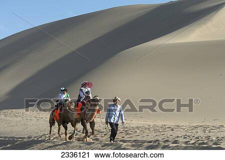 Stock Image - Tourists Riding On Camels At Singing Sand Mountain; Jiuquan, Gansu, China. Fotosearch