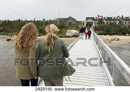 Stock Photography - Tourists walking on dock towards buildings on shore, Gros Morne National Park; Newfoundland, Canada. Fotosearch