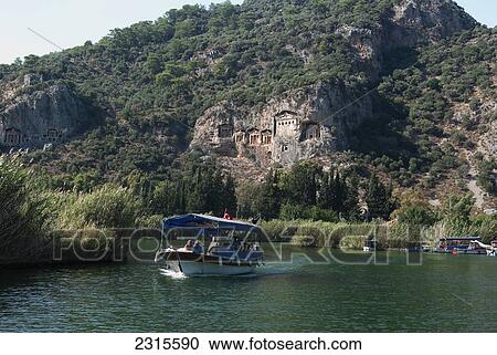 Stock Image - Turkey, Dalyan, Small tourist boat passing Lycian style Kings Tombs on Dalyan River. Fotosearch