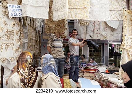 Stock Image - Turkey, Table Cloth's And Bedspreads On Display At Carsamba Pazari; Istanbul, Shoppers Looking At Linens. Fotosearch
