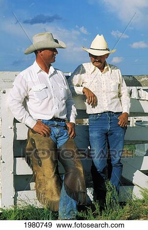 Stock Photo - Two Cowboys Leaning Against /Nwhite Fence In Bright Sunlight, Wearing Long Sleeve Shirts, Jeans And Cowboy Hats.. Fotosearch