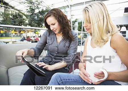 Stock Image - Two friends looking at a tablet while having coffee in a mall; St. Albert, Alberta, Canada. Fotosearch