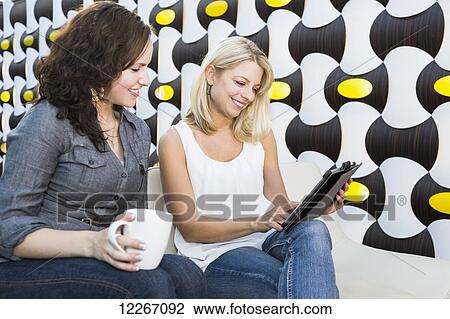 Stock Image - Two friends looking at a tablet while having coffee in a mall; St. Albert, Alberta, Canada. Fotosearch