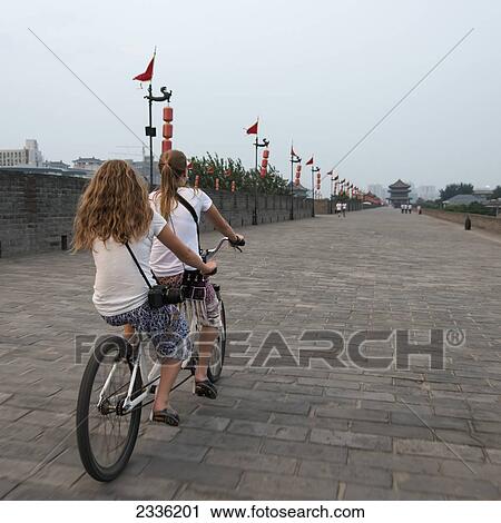 Stock Image - Two Girls Touring On A Tandem Bicycle Along Lianhu Ancient City Wall; Xi'an, Shaanxi, China. Fotosearch