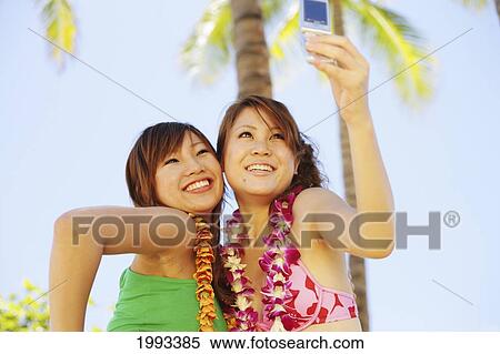 Stock Photography - Two Japanese Girls Take A Picture Of Themselves With A Camera Phone, Palm Trees Behind. Fotosearch