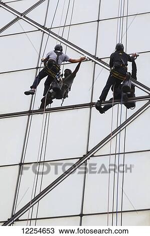 Stock Image - Two men in harnesses cleaning exterior of windows on modern building; Portugal. Fotosearch