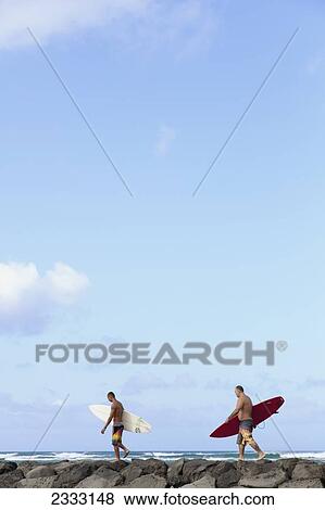 Stock Photo - Two men walk along rock wall with surfboards; Honolulu, Oahu, Hawaii, United States of America. Fotosearch