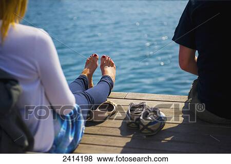 Stock Photo - Two people sitting on a wooden dock at the water's edge; Barcelona, Spain. Fotosearch