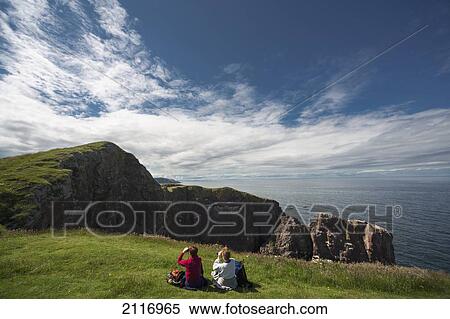 Stock Photography - Two People Sitting On The Grass Looking Through Binoculars At The Rocky Promontory At St. Abb's Head; Scottish Borders Scotland. Fotosearch