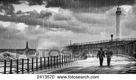 Two people walking along the water's edge in the rain with waves crashing into the shore and a lighthouse in the background; Sunderland, Tyne and Wear, England View Large Photo Image Stock Image - Two people walking along the water's edge in the rain with waves crashing into the shore and a lighthouse in the background; Sunderland, Tyne and Wear, England. Fotosearch
