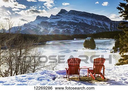 Stock Photograph - Two red chairs on snow covered ridge overlooking frozen lake with snow covered mountain in the background with blue sky and clouds; Banff, Alberta, Canada. Fotosearch