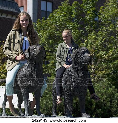 Stock Image - Two teenage girls pose sitting atop dog statues; St. John's, Newfoundland, Canada. Fotosearch