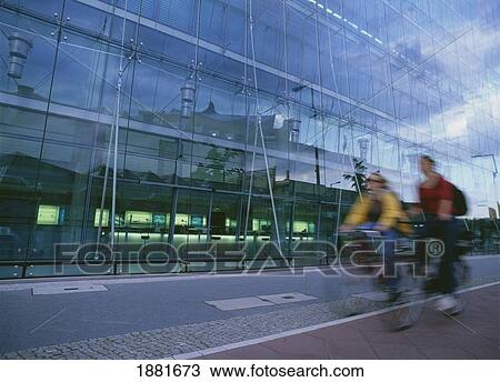 Stock Image - Two Women Cycling Past Modern Architecture, Blurred Motion. Fotosearch
