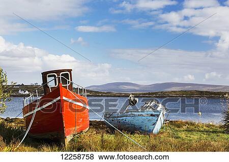 Two wooden boats, red and blue, on grassy shore with bay in background with clouds and blue sky; Roundstone, County Galway, Ireland View Large Photo Image Stock Photo - Two wooden boats, red and blue, on grassy shore with bay in background with clouds and blue sky; Roundstone, County Galway, Ireland. Fotosearch