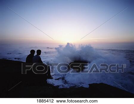 Stock Image - Two Young Boys Watching Waves Coming Crashing Into Rocks And The Sea Walls Of Essaouira At Dusk. Fotosearch