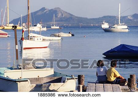 Stock Photograph - Two Young Children Sitting On A Dock In Port De Mollenca. Fotosearch