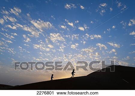 Stock Image - Two young girls running up sand dune at dusk; Dubai, United Arab Emirates. Fotosearch