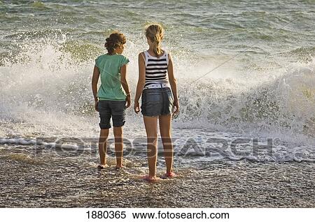 Two Young Girls Standing At The Beach With Their Feet In The Water. View Large Photo Image Stock Photography - Two Young Girls Standing At The Beach With Their Feet In The Water.. Fotosearch