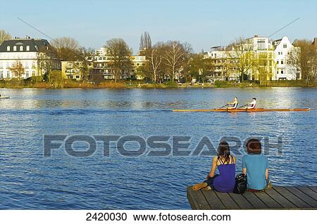 Stock Image - Two young people sit on a wooden dock along the river; Hamburg, Germany. Fotosearch