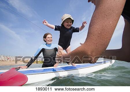 Stock Image - Two Young Women And One Year Old Toddler Playing With A Standup Paddleboard Off The Beachfront At Hayling Island. Fotosearch