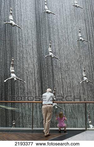 Stock Photo - UAE, Man and girl looking at large installation of waterfall and divers in Dubai Mall shopping centre; Dubai. Fotosearch