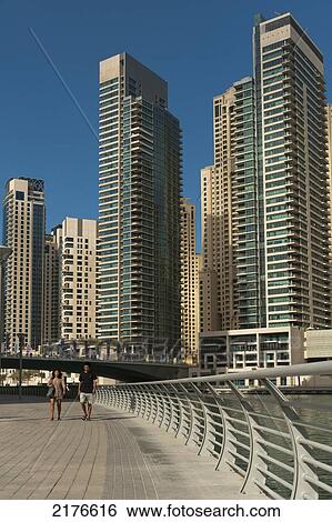 Stock Photograph - UAE, People walking through Dubai Marina area with large residential blocks of flats behind; Dubai. Fotosearch