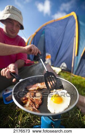 Stock Image - UK, Dorset, Man cooking fried breakfast of bacon and egg beside tent; Lulworth. Fotosearch