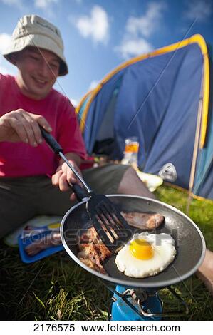 Stock Photography - UK, Dorset, Man cooking fried breakfast of bacon and egg beside tent; Lulworth. Fotosearch