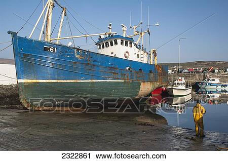 Stock Image - UK, Ireland, County Kerry, Iveragh Peninsula, Portmagee, Fisherman standing on wet shore beside large fishing vessel. Fotosearch