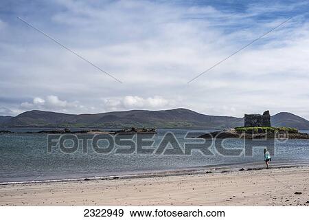 Stock Photo - UK, Ireland, County Kerry, Woman walking on beach with Ballinskelligs Castle in background. Fotosearch