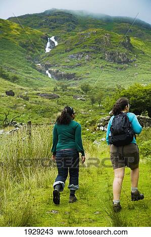 Stock Image - UK, North Wales, Snowdonia National Park, Waterfall Cascades in Distance; Nantgwynant, Two hikers on trail. Fotosearch