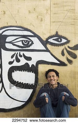 Stock Image - UK, Teenage boy sitting in front of graffiti wall of cubist style face; Hastings. Fotosearch