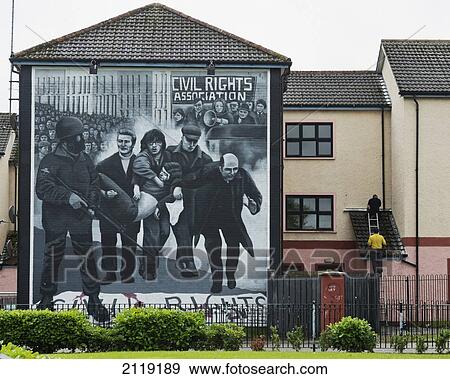 Stock Photo - United Kingdom, Northern Ireland, County Londonderry, Two workmen on ladder working next to Bloody Sunday mural in Rossville Street of Bogside Area; Derry. Fotosearch