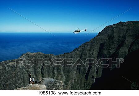 Stock Photo - USA, Hawaii Islands, Kauai, Helicopter in air; Kalalau Lookout, Hikers overlooking valley and ocean. Fotosearch