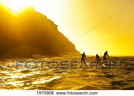 Stock Photo - USA, Hawaii Islands, Oahu, Three Stand Up Paddlers Passing Spitting Caves At Sunrise; Portlock. Fotosearch