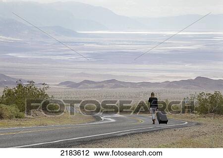 Stock Image - Usa, Nevada Border, California, Route 374; Death Valley National Park, Highway 374, Young Woman Walking On Deserted Road In Desert. Fotosearch