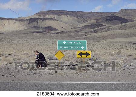 Picture - Usa, Route 136 Near Death Valley; California, Young Woman Sitting By Lone Pine And Furnace Creek Signs On Highway 136. Fotosearch