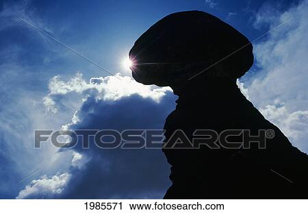 Stock Image - Utah, Arches National Park, Balanced Rock Silhouetted Against Sunlight, Clouds In Blue Sky.. Fotosearch